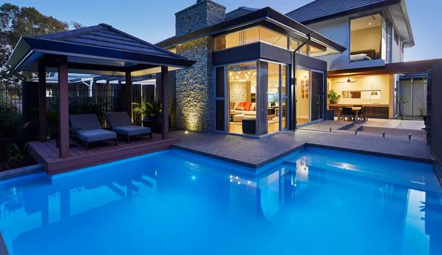 Custom home swimming pool and alfresco area featuring timber decking, expansive glazing and outdoor lighting at dusk.